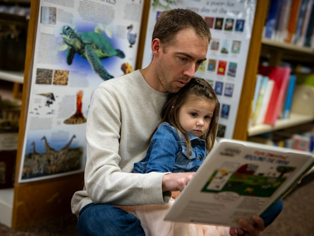 Ein Vater liest seiner kleinen Tochter in einer Bibliothek konzentriert aus einem Buch vor – gelebte Sprachförderung im Alltag.
